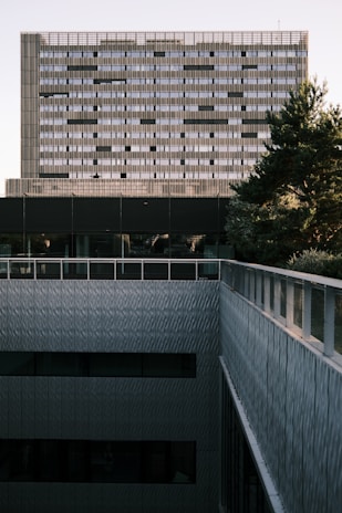 A modern architectural facade featuring a grid of rectangular windows on a high-rise building. In the foreground, there is a terrace with metal railings, while a tree partially obscures the view on the right. The building has a geometric and minimalist design, giving a sense of structure and symmetry.