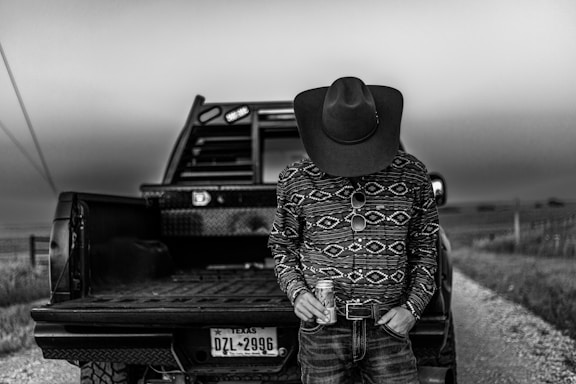 A person wearing a patterned long-sleeve shirt, jeans, and a cowboy hat stands in front of an open pickup truck on a rural road. The head is bowed, obscured by the hat, and the person holds a can in one hand. The scene is set in black and white, with a license plate from Texas visible on the truck.