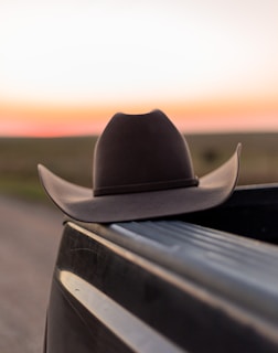 A cowboy hat rests on the rim of a truck's bed, with an expansive blurred landscape and soft hues of a sunset in the background.