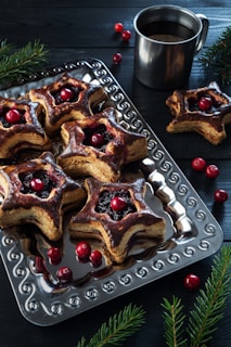 A metal tray holds star-shaped pastries with a glossy brown crust, filled with dark fruit and topped with red berries. Accompanying the pastries is a metallic mug filled with a dark beverage. The scene is adorned with scattered red cranberries and sprigs of evergreen branches, set against a dark wooden background.