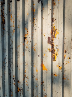Corrugated metal surface exhibiting signs of weathering and rust, with patches of peeling yellow and brown paint against a backdrop of gray metal.