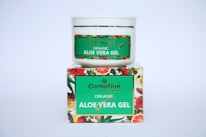 A white container with a green label for 'Carnation Organic Aloe Vera Gel' is placed on top of a colorful box featuring images of various fruits and flowers.
