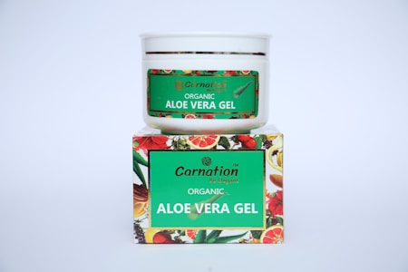 A white container with a green label for 'Carnation Organic Aloe Vera Gel' is placed on top of a colorful box featuring images of various fruits and flowers.