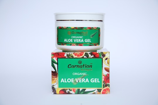 A white container with a green label for 'Carnation Organic Aloe Vera Gel' is placed on top of a colorful box featuring images of various fruits and flowers.