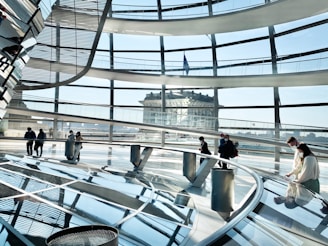 An interior view of a modern architectural structure with a glass dome, featuring curved glass panels and a reflective floor. Several people are walking and observing the surroundings, wearing face masks. In the background, an older classical building is visible through the glass.