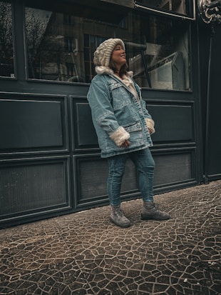 A person wearing a warm outfit, including a cozy denim coat with fur trim, jeans, and boots. They are standing on a patterned stone floor in front of a dark-colored paneled building, looking content and relaxed.