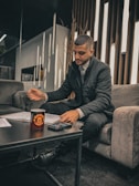 A person seated on a gray sofa is writing with a pen on papers spread over a dark coffee table. A cup with a logo and a calculator are also on the table. The setting appears to be a modern, well-lit lounge with narrow vertical wooden strips on the wall behind.
