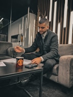 A person seated on a gray sofa is writing with a pen on papers spread over a dark coffee table. A cup with a logo and a calculator are also on the table. The setting appears to be a modern, well-lit lounge with narrow vertical wooden strips on the wall behind.