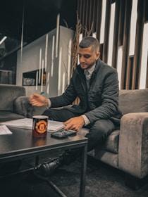 A person seated on a gray sofa is writing with a pen on papers spread over a dark coffee table. A cup with a logo and a calculator are also on the table. The setting appears to be a modern, well-lit lounge with narrow vertical wooden strips on the wall behind.