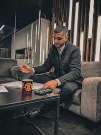 A person seated on a gray sofa is writing with a pen on papers spread over a dark coffee table. A cup with a logo and a calculator are also on the table. The setting appears to be a modern, well-lit lounge with narrow vertical wooden strips on the wall behind.