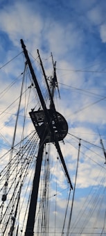 A ship's mast with a complex network of ropes and rigging against a backdrop of a partly cloudy sky.