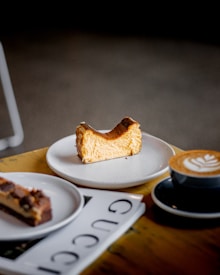 A slice of Basque cheesecake is placed on a white plate, accompanied by a cup of cappuccino with latte art. Nearby, a piece of chocolate dessert rests on another plate, all arranged on a wooden table alongside a magazine.