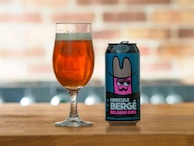 A can of 'Hercule Bergé Belgian DIPA' beer with a colorful label featuring an illustration of a character wearing a wide-brimmed hat is placed on a wooden surface next to a glass filled with amber-colored beer. The background is softly blurred, showcasing a brick pattern.