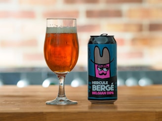 A can of 'Hercule Bergé Belgian DIPA' beer with a colorful label featuring an illustration of a character wearing a wide-brimmed hat is placed on a wooden surface next to a glass filled with amber-colored beer. The background is softly blurred, showcasing a brick pattern.