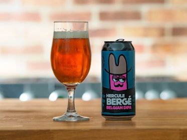 A can of 'Hercule Bergé Belgian DIPA' beer with a colorful label featuring an illustration of a character wearing a wide-brimmed hat is placed on a wooden surface next to a glass filled with amber-colored beer. The background is softly blurred, showcasing a brick pattern.