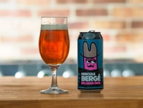 A can of 'Hercule Bergé Belgian DIPA' beer with a colorful label featuring an illustration of a character wearing a wide-brimmed hat is placed on a wooden surface next to a glass filled with amber-colored beer. The background is softly blurred, showcasing a brick pattern.