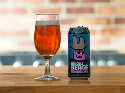 A can of 'Hercule Bergé Belgian DIPA' beer with a colorful label featuring an illustration of a character wearing a wide-brimmed hat is placed on a wooden surface next to a glass filled with amber-colored beer. The background is softly blurred, showcasing a brick pattern.