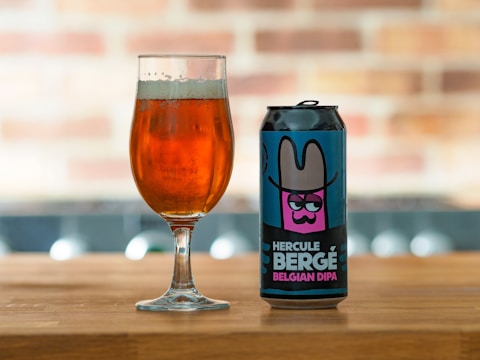 A can of 'Hercule Bergé Belgian DIPA' beer with a colorful label featuring an illustration of a character wearing a wide-brimmed hat is placed on a wooden surface next to a glass filled with amber-colored beer. The background is softly blurred, showcasing a brick pattern.