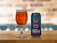 A can of 'Hercule Bergé Belgian DIPA' beer with a colorful label featuring an illustration of a character wearing a wide-brimmed hat is placed on a wooden surface next to a glass filled with amber-colored beer. The background is softly blurred, showcasing a brick pattern.