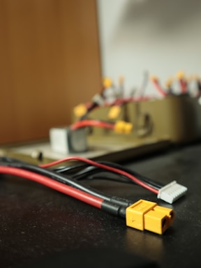 A close-up view of several electrical connectors and cables on a dark surface. The focus is on a yellow connector with a plus and minus sign, attached to red and black wires. In the background, other wires and connectors are visible, but they are blurry.