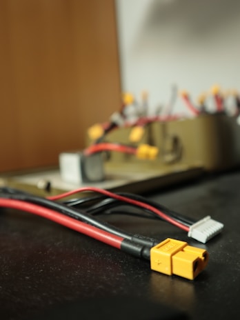 A close-up view of several electrical connectors and cables on a dark surface. The focus is on a yellow connector with a plus and minus sign, attached to red and black wires. In the background, other wires and connectors are visible, but they are blurry.