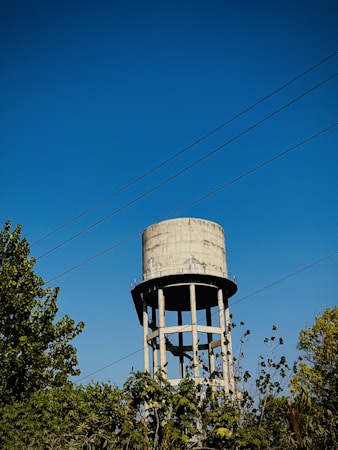 A tall, cylindrical water tower stands against a clear blue sky. Several power lines stretch horizontally across the sky, and lush greenery surrounds the base of the tower.