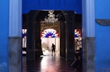A person is mopping the floor inside an open architectural doorway. The interior features a chandelier and stained glass windows with geometric patterns in red and blue, while the entrance is framed by blue-painted walls.