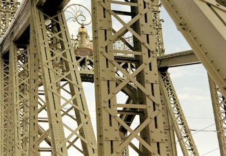 Intricate metal framework featuring a large bell positioned centrally, with ornate scrollwork and a visible sky in the background. The structure's crisscrossing beams suggest an industrial or architectural setting.