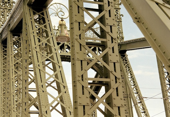 Intricate metal framework featuring a large bell positioned centrally, with ornate scrollwork and a visible sky in the background. The structure's crisscrossing beams suggest an industrial or architectural setting.