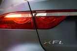 Close-up view of a car's taillight with illuminated red lights, featuring the silver letters 'XFL' on the vehicle's body. The car has a metallic grey finish and the light is partially covered with a chrome strip.