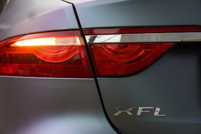 Close-up view of a car's taillight with illuminated red lights, featuring the silver letters 'XFL' on the vehicle's body. The car has a metallic grey finish and the light is partially covered with a chrome strip.