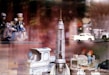 A collection of vintage toys featuring a variety of vehicles including a space rocket, motorcycles, and trucks. Some toys have figurines, such as a motorcycle with riders and a truck with two people inside. The toys appear to be displayed on glass shelves with reflections from the glass.