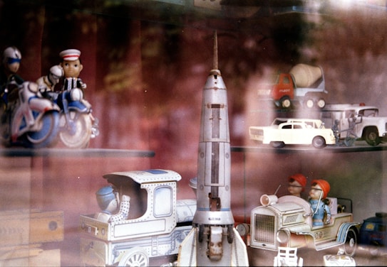 A collection of vintage toys featuring a variety of vehicles including a space rocket, motorcycles, and trucks. Some toys have figurines, such as a motorcycle with riders and a truck with two people inside. The toys appear to be displayed on glass shelves with reflections from the glass.