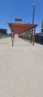 A wooden pergola structure extends over a concrete walkway with tall buildings in the background and clear blue skies above. Metal lampposts are positioned along the path, providing lighting. Trees and greenery can be seen on the left side of the image, adding a touch of nature to the urban setting.