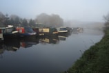 Several narrowboats are moored along a calm, foggy canal with trees and vegetation on one side. Reflections of the boats are visible in the water. The atmosphere appears quiet and serene, with a misty background limiting visibility.