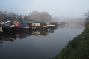 Several narrowboats are moored along a calm, foggy canal with trees and vegetation on one side. Reflections of the boats are visible in the water. The atmosphere appears quiet and serene, with a misty background limiting visibility.
