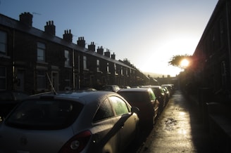 A row of terraced houses lines the street under a clear sky. Parked cars are tightly arranged along the wet pavement, reflecting the sunlight. The bright sun is low in the sky, casting long shadows and a warm glow over the scene.