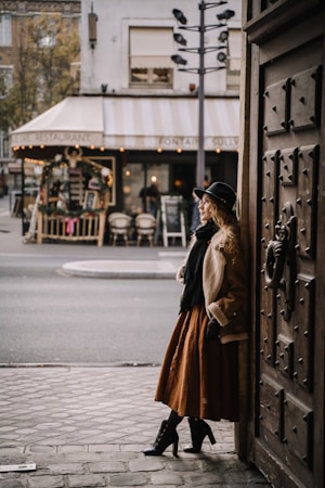 A woman in stylish, vintage-inspired attire leans against a large wooden door. She wears a long brown skirt, a coat, a black scarf, and a black hat, looking contemplative. In the background, a cozy restaurant with a striped awning and outdoor seating is visible, creating a quaint and charming urban scene.