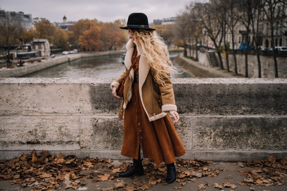 a woman in a long coat and hat standing on a stone ledge