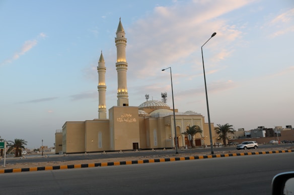 A large, elegant mosque with two tall minarets and a central dome against a partly cloudy sky. The mosque is beige-colored with intricate architectural details and Arabic script on its facade. Palm trees and a paved road with vehicles are visible in the foreground.