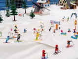 A miniature diorama depicting a lively ski resort with various cartoonish figures engaged in skiing and snow activities. Small buildings typical of a mountain resort and evergreen trees surround the scene, while colorful figurines are found skiing, playing, and interacting on the snow.