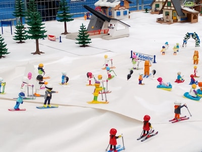 A miniature diorama depicting a lively ski resort with various cartoonish figures engaged in skiing and snow activities. Small buildings typical of a mountain resort and evergreen trees surround the scene, while colorful figurines are found skiing, playing, and interacting on the snow.