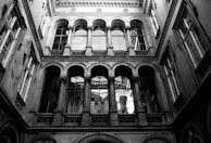 A black and white photograph of an ornate architectural structure featuring arched windows and intricate stonework. The perspective looks upwards from a courtyard, capturing the symmetry and classical elegance of the building.