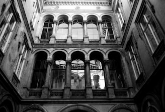 A black and white photograph of an ornate architectural structure featuring arched windows and intricate stonework. The perspective looks upwards from a courtyard, capturing the symmetry and classical elegance of the building.