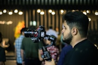 A person is holding a professional camera mounted on a stabilizing gimbal, standing amidst a dimly lit setting with bright lights blurred in the background. Several people are present, though out of focus, creating a lively atmosphere.