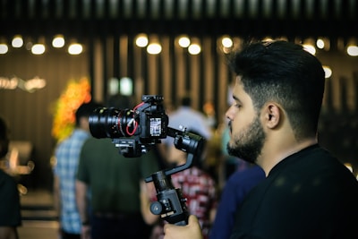 A person is holding a professional camera mounted on a stabilizing gimbal, standing amidst a dimly lit setting with bright lights blurred in the background. Several people are present, though out of focus, creating a lively atmosphere.
