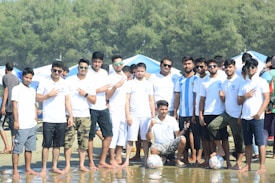 A group of men wearing casual clothing, mostly white shirts, are gathered on a sandy area near water. Some are posing with fingers forming peace signs, and two soccer balls are visible at their feet. Trees and tents are visible in the background.