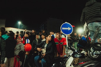 A group of people gathered, some sitting on scooters, with a prominent right-turn road sign and streetlights illuminating the scene. The crowd appears to be diverse, with individuals dressed in various styles. The image captures a moment of anticipation or curiosity, as many people look in the same direction.