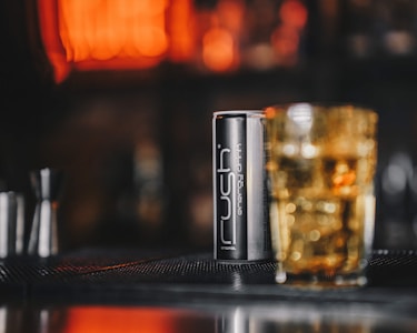 A can of Rush energy drink is placed on a bar mat, beside a glass filled with a golden-colored drink. The background is softly blurred, featuring warm, orange tones, suggesting a cozy bar or nightclub setting.