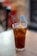 A glass filled with iced tea is placed on a wooden table. The drink is topped with a wedge of lemon and contains a silver spoon and a blue straw.